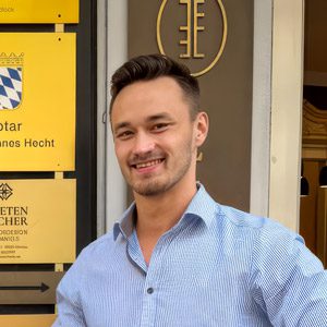 Junger Mann mit braunen Haaren lächelt vor einem Büro.