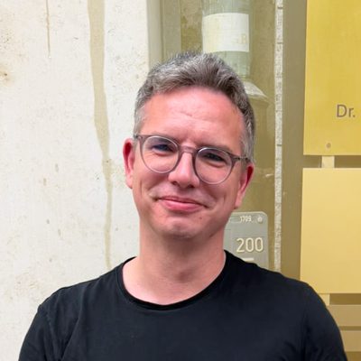 Mann mit grauen Haaren und Brille lächelt vor einer Wand mit Schildern.