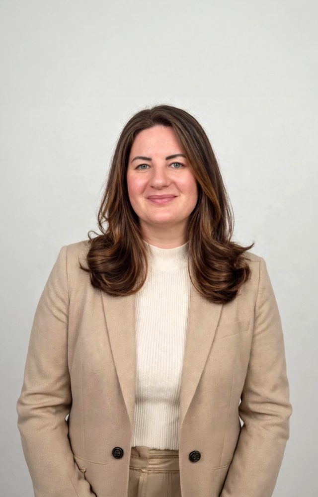 Porträt einer Person mit langen Haaren in einem hellen Blazer auf neutralem Hintergrund.